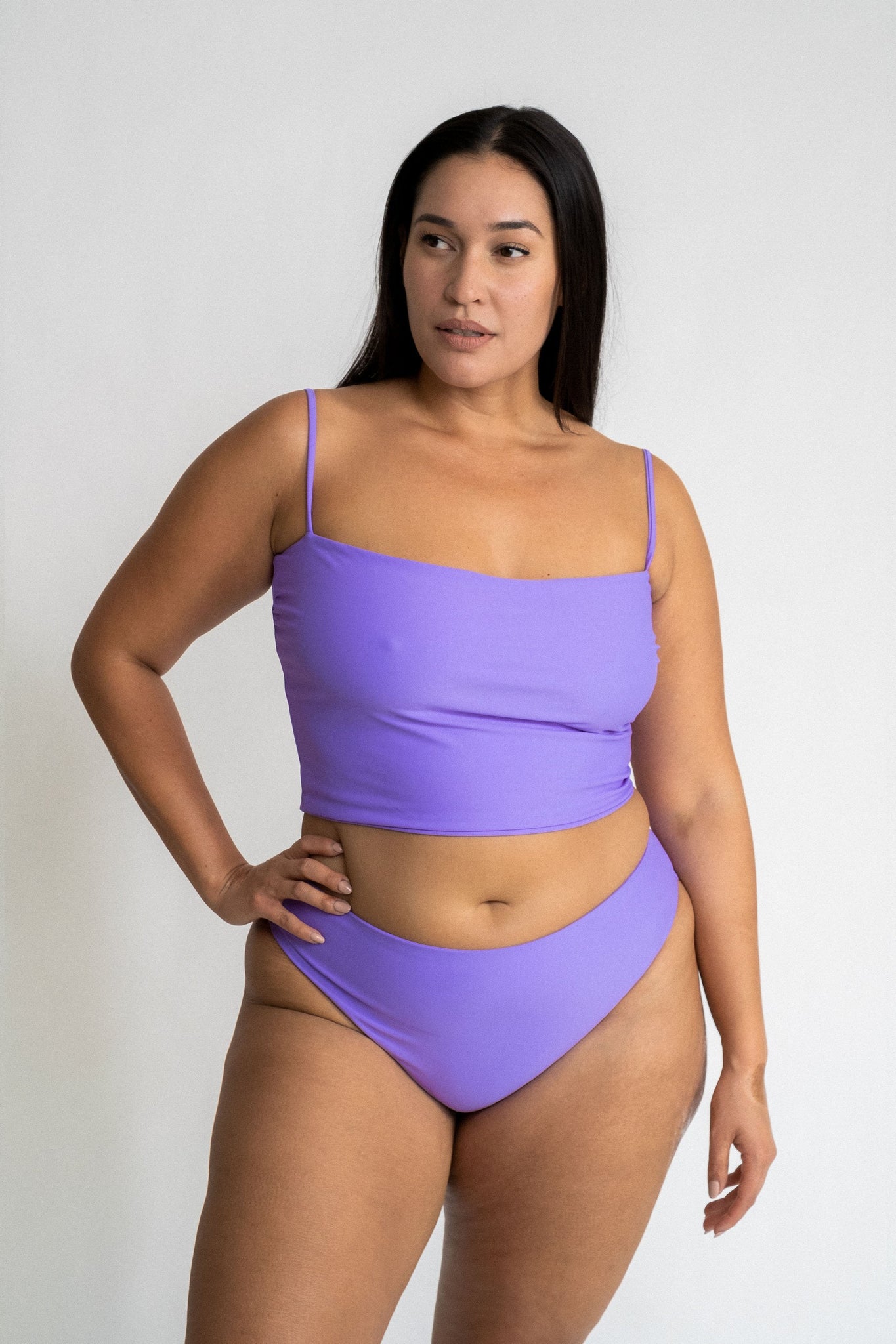 A woman standing with her hand on her hip looking to the side wearing bright lavender high cut bikini bottoms and a matching bright lavender tankini.
