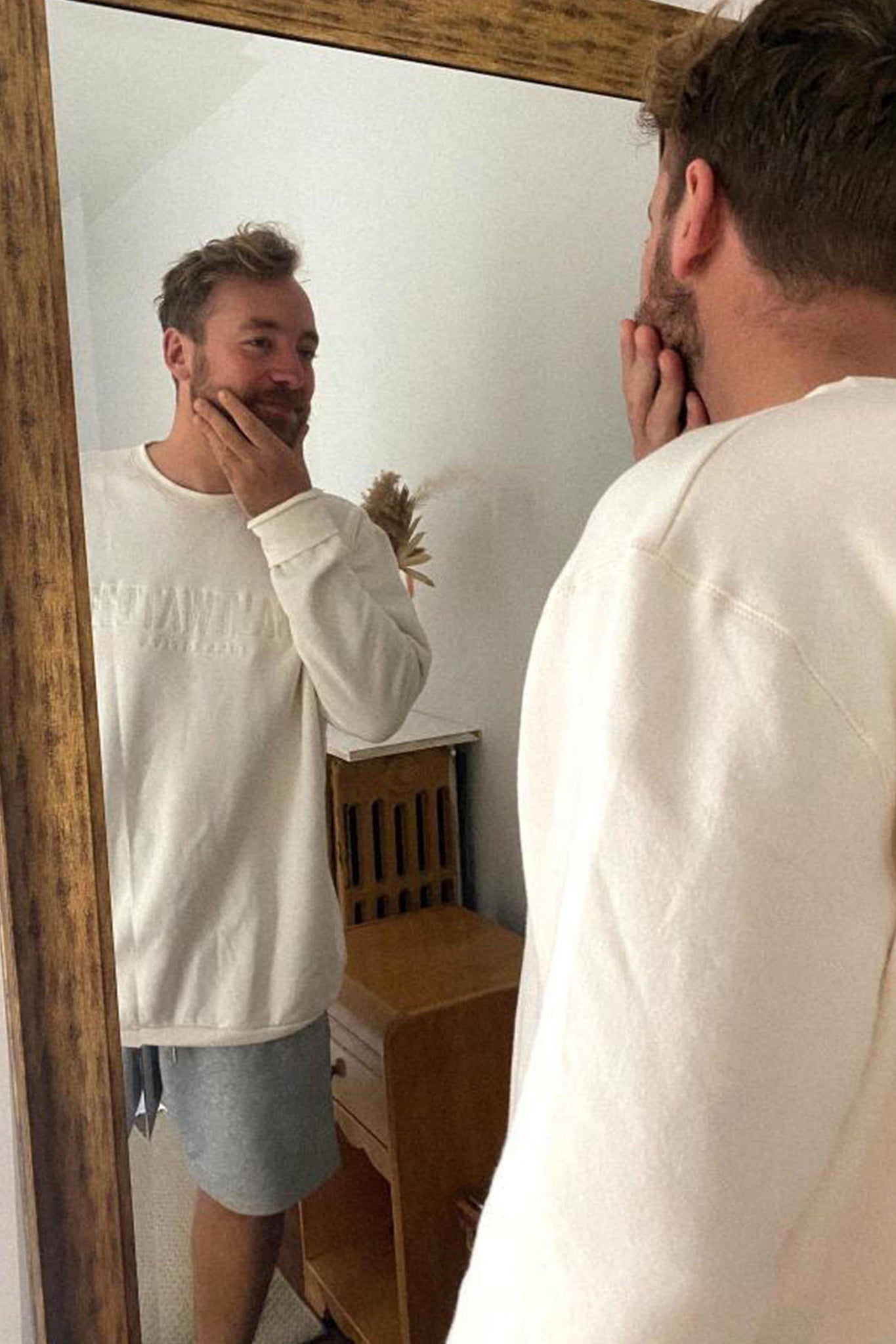 A man looking in the mirror with his hand on his chin wearing an oversized white sweatshirt and grey sweatshorts. 