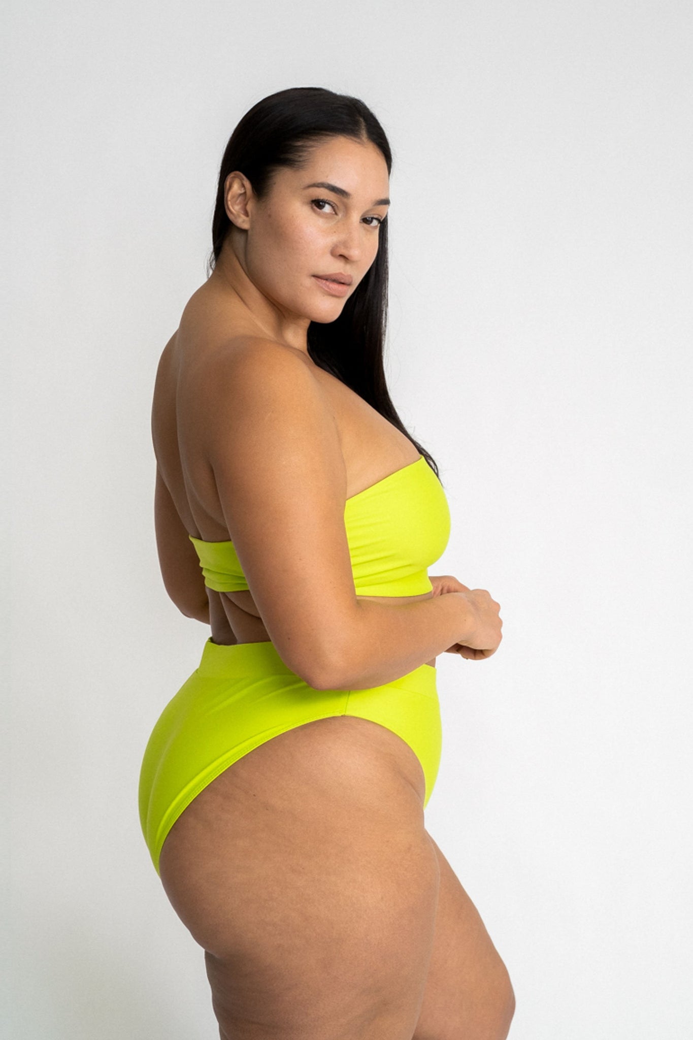 A woman looking over her shoulder wearing bright neon green high waisted bikini bottoms with a matching bright neon green strapless bandeau bikini top.