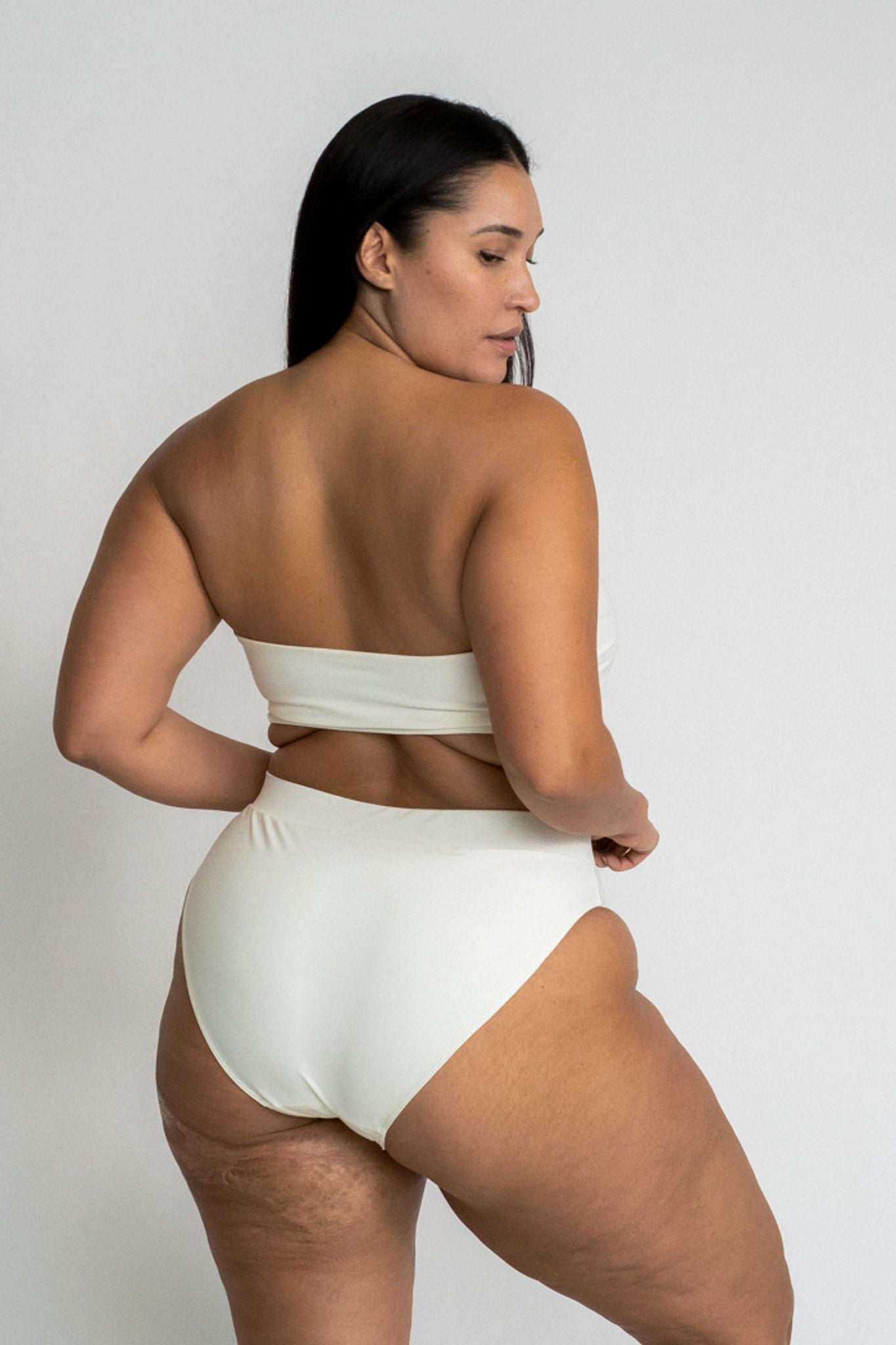 A woman standing in front of a white wall looking over her shoulder wearing white high waisted bikini bottoms with a matching white bandeau strapless bikini top.