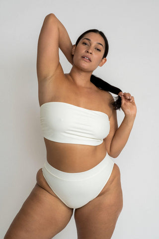 A woman standing holding her hair wearing white high waisted bikini bottoms with a matching white bandeau strapless top.
