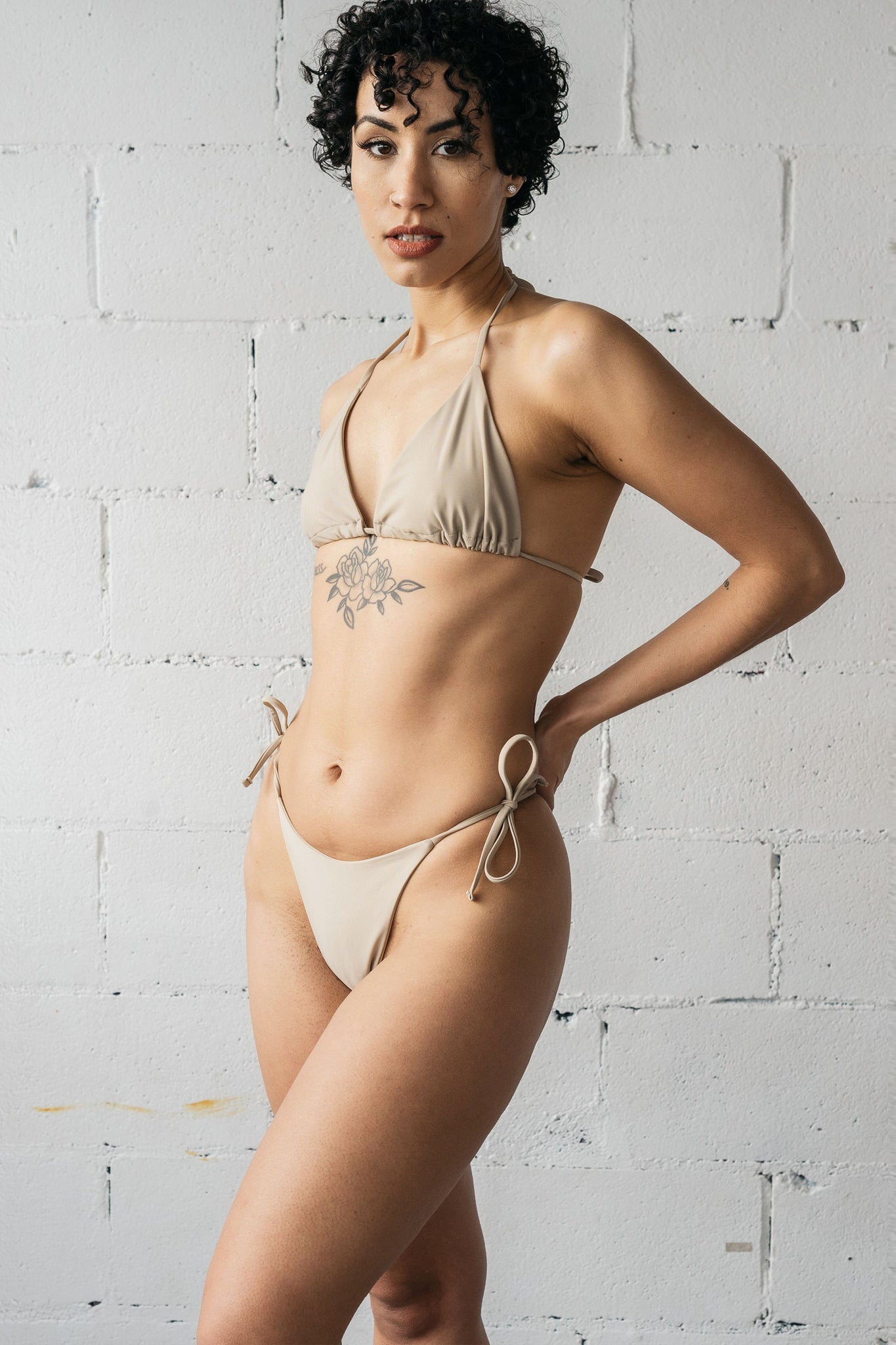 A woman standing with one leg placed in front of the other wearing nude adjustable string bikini bottoms with a matching nude string triangle bikini top.