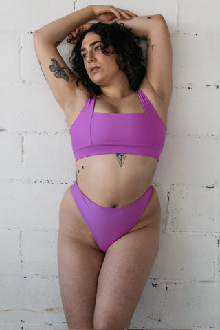 A woman leaning against a white wall with both arms above her head wearing bright pink high waisted bikini bottoms and a matching bright pink bikini top with a square neckline.