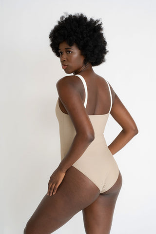The back of a woman with her arms on her thighs looking over her shoulder wearing a nude one piece swimsuit with thick white spaghetti straps.