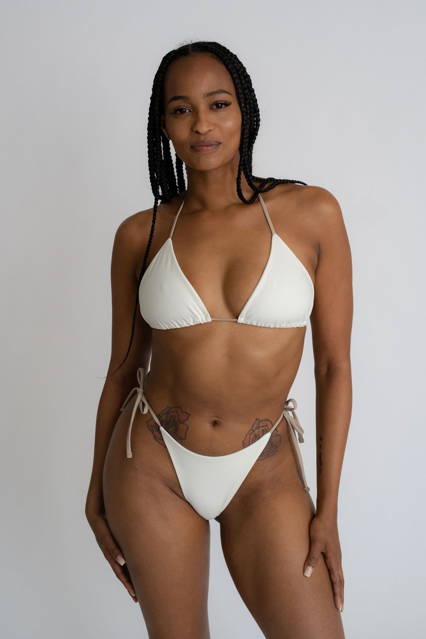 A woman standing with her hands hung by her side wearing white triangle bikini bottoms with nude adjustable strings with a matching white triangle bikini top and nude adjustable straps.