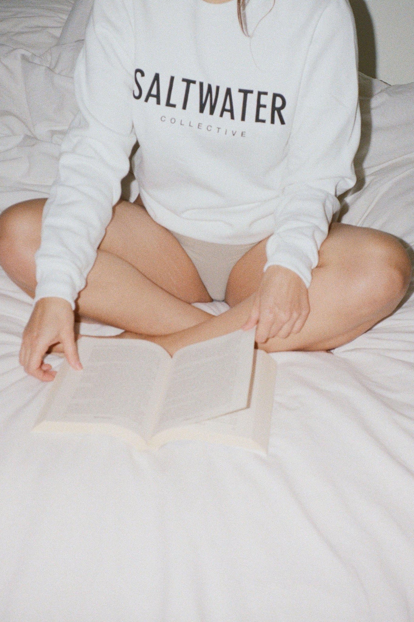 A close up of a woman holding a book while sitting on a bed wearing a white crewneck sweatshirt with a SALTWATER logo printed across the chest in black.