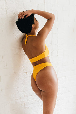The back of a woman leaning against a white wall wearing high cut yellow bikini bottoms wearing a matching yellow bikini top.