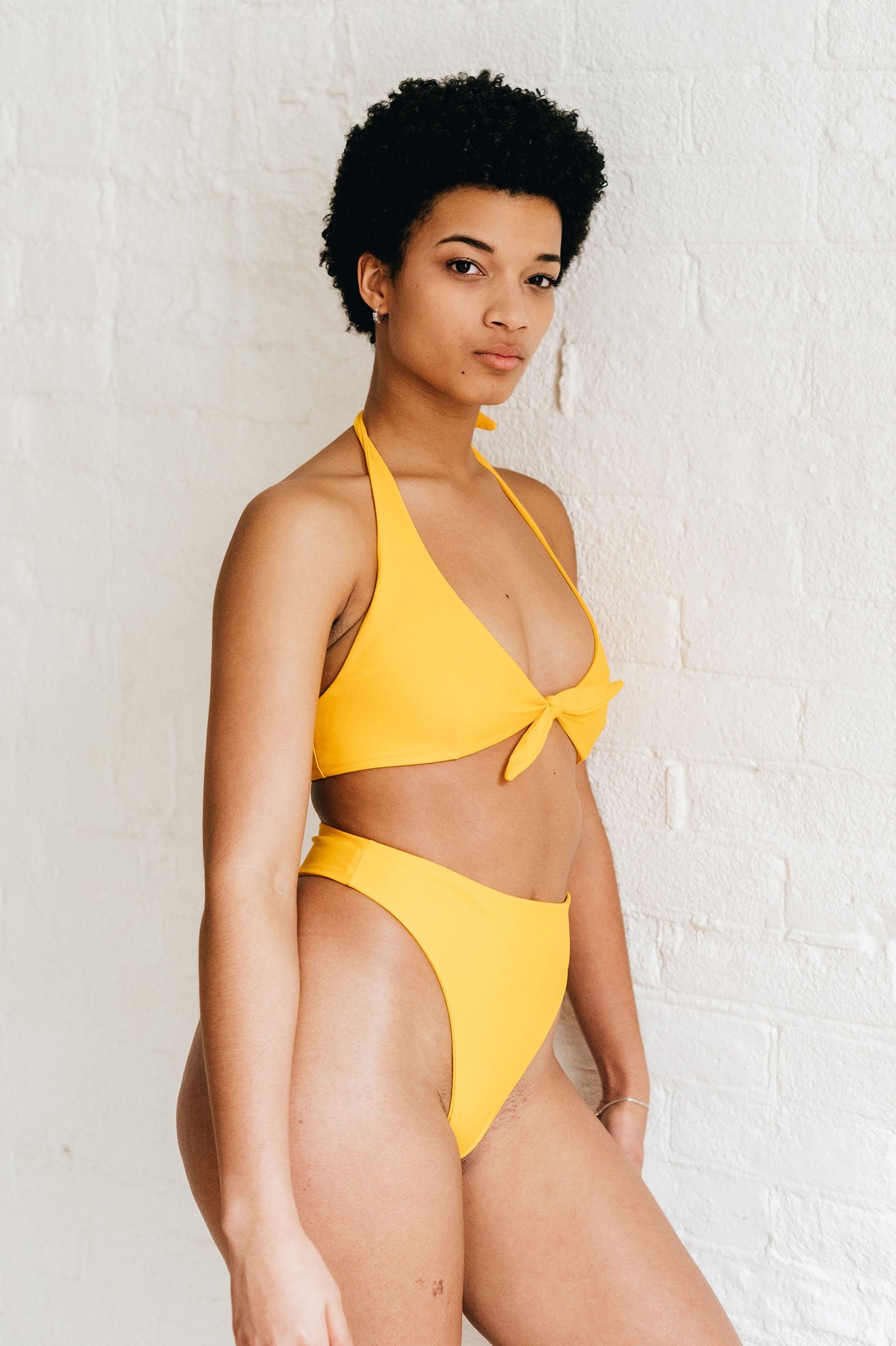 A woman standing to the side wearing high cut yellow bikini bottoms with a matching tie front yellow bikini top.