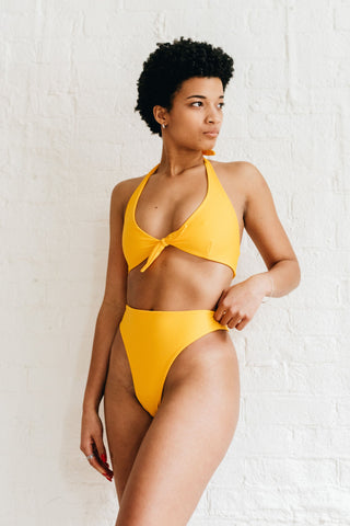 A woman standing with one hand on her hip looking away wearing high cut yellow bikini bottoms with a matching tie front yellow bikini top.