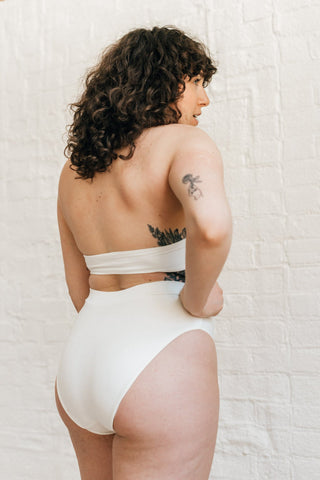 The back of a woman standing with one hand on her hip wearing high waisted white bikini bottoms with full coverage and a matching white bikini top.