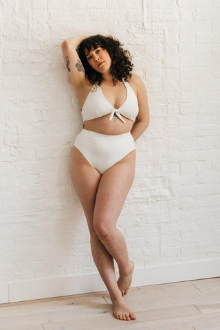 A woman leaning against a white wall wearing high waisted white bikini bottoms with full coverage and a matching white tie front bikini top.