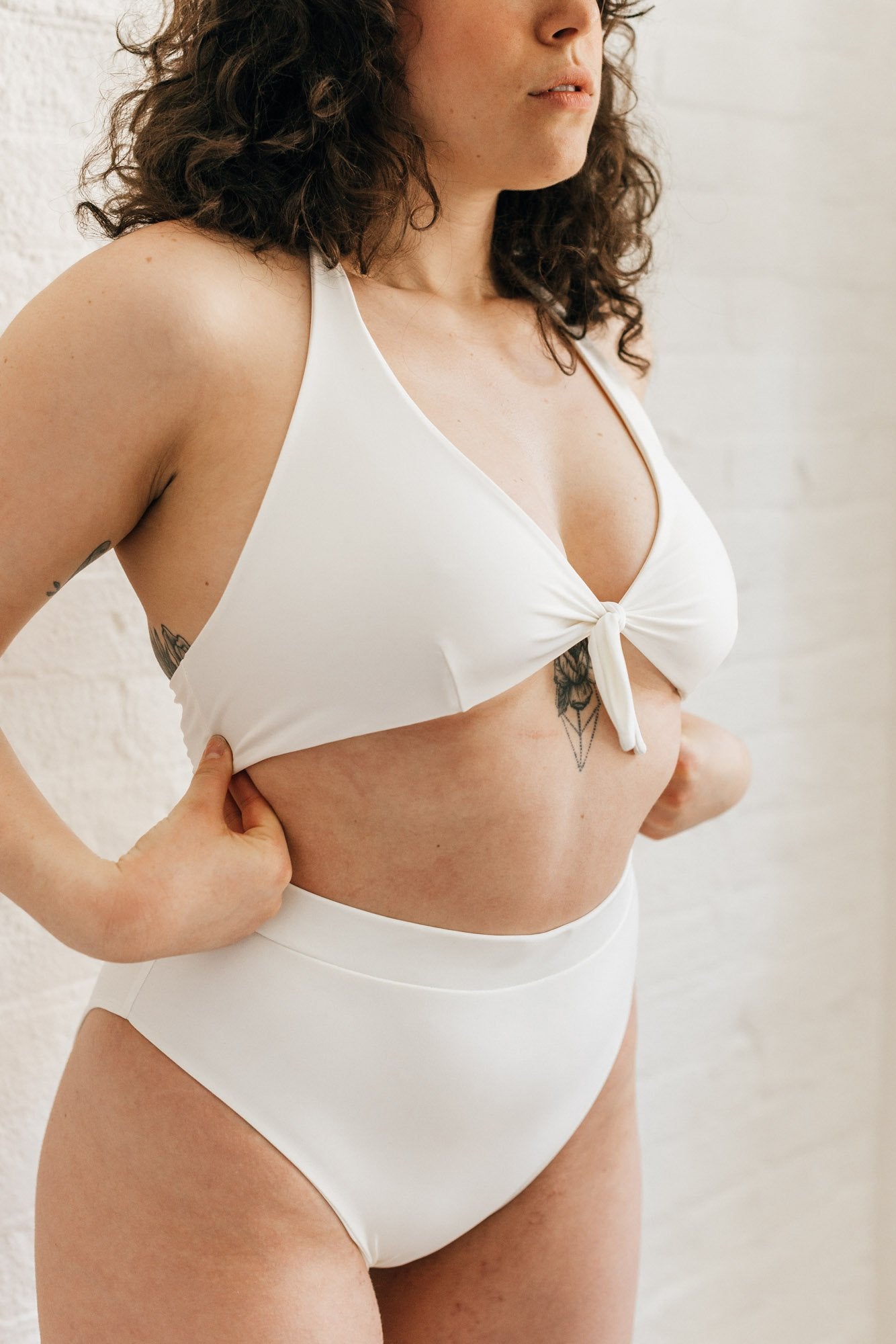 A woman standing to the side wearing high waisted white bikini bottoms with full coverage and a matching white tie front bikini top.