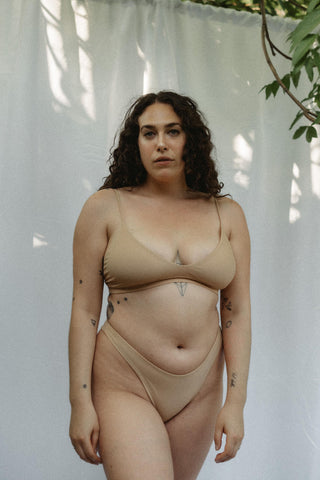 Woman standing against a white backdrop wearing a beige swimsuit with high cut bottoms and a strappy top