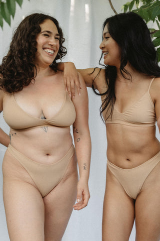 Two women laughing and wearing matching beige swimsuits featuring high cut bottoms and spaghetti strap tops