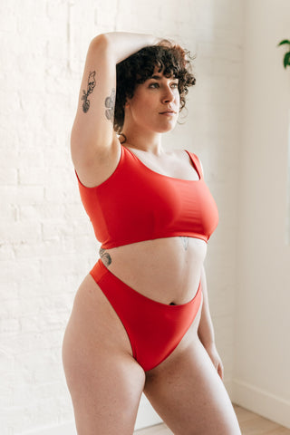 A woman standing with one arm above her head wearing a red bikini that features full coverage thick straps and high waisted bottoms