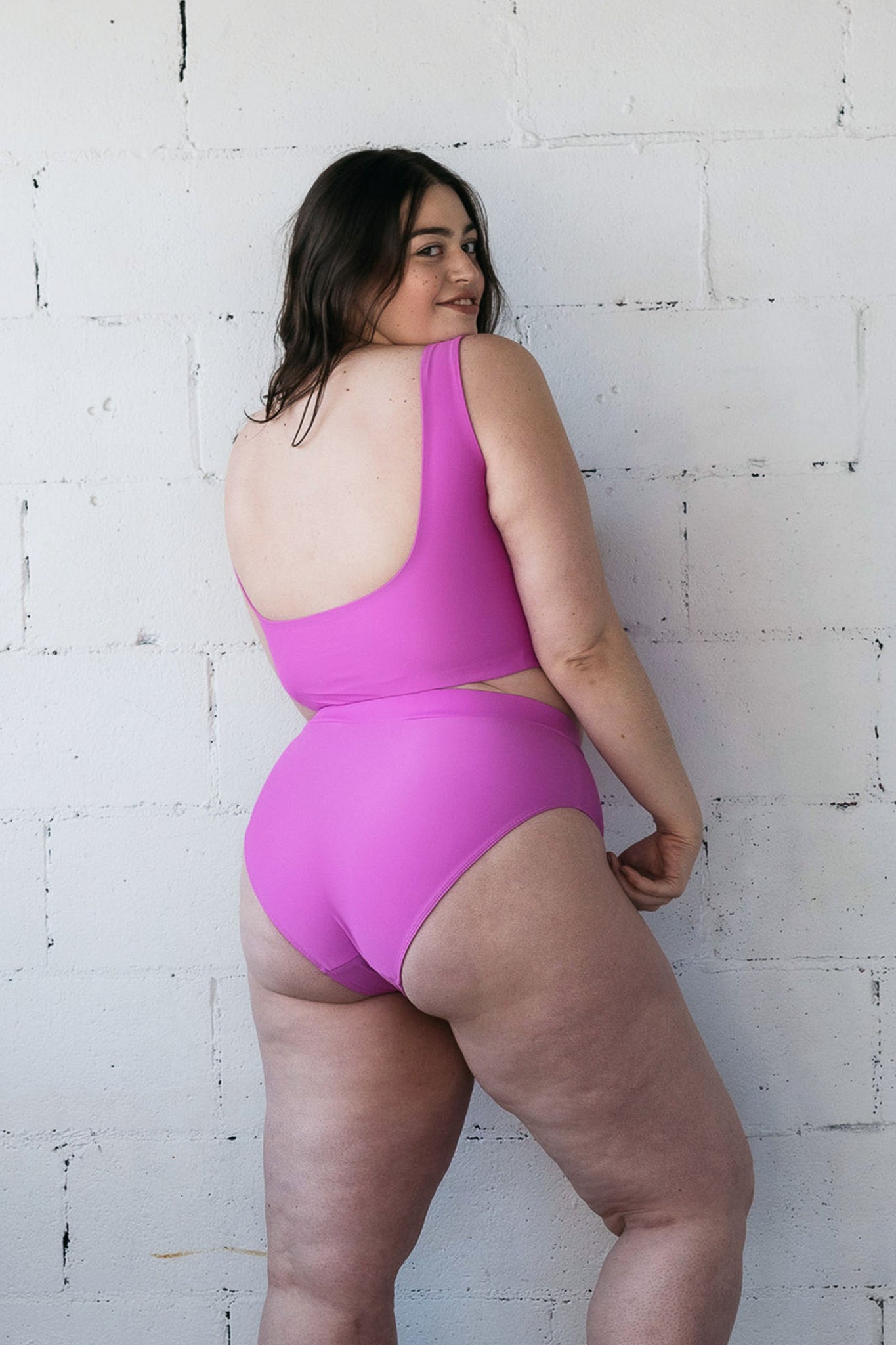 A woman looking over her shoulder and smiling wearing a bright purple bikini with high waisted bottoms.