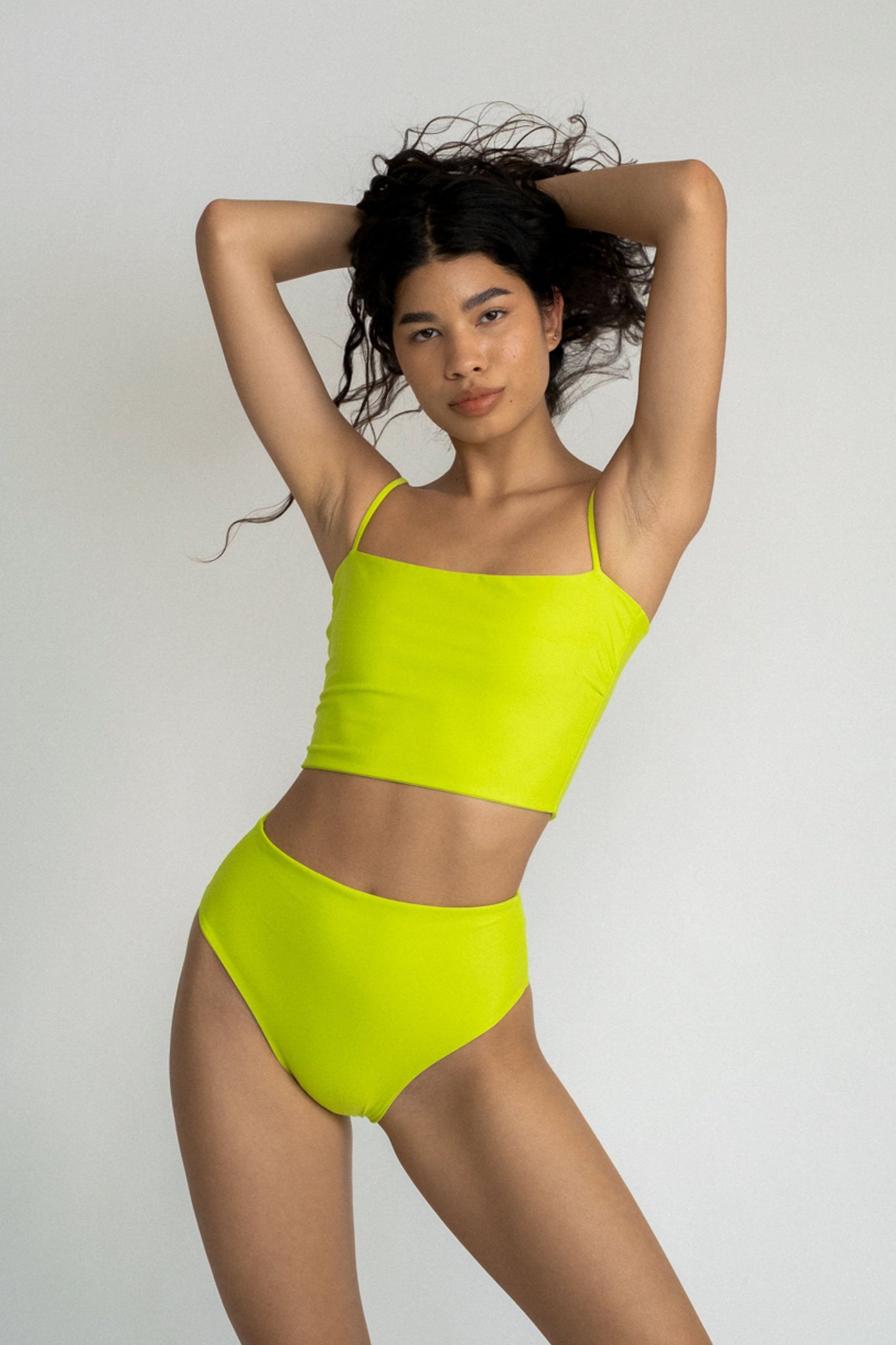 A woman standing with her hands in her hair wearing a bright neon green tankini top with matching bright neon green high waisted bikini bottoms.