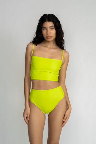 A woman standing with her hands by her side wearing a bright neon green tankini top and matching bright neon green high waisted bikini bottoms.