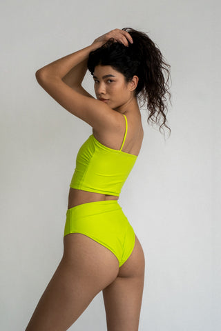 A woman standing and looking over her shoulder with her hands in her hair wearing a bright neon green high waisted bikini bottoms with a matching bright neon green tankini top.