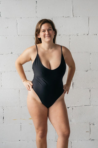 A woman standing with her hands on her hips wearing a black one-piece swimsuit featuring a low neckline and spaghetti straps.