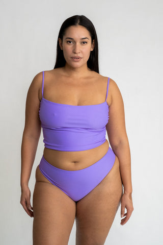 A woman standing with her arms by her side wearing bright lavender high-waisted bikini bottoms and a matching bright lavender tankini top.