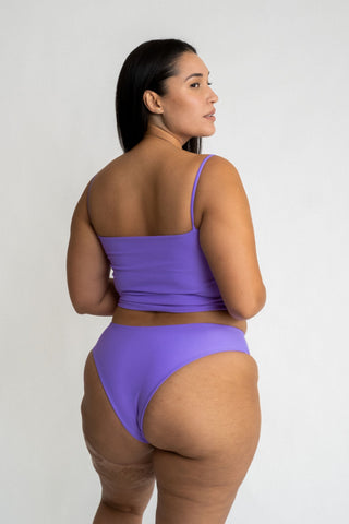 The back of a woman looking to the side with  her arms crossed wearing high cut bright purple bikini bottoms with a matching bright purple tankini bikini top.