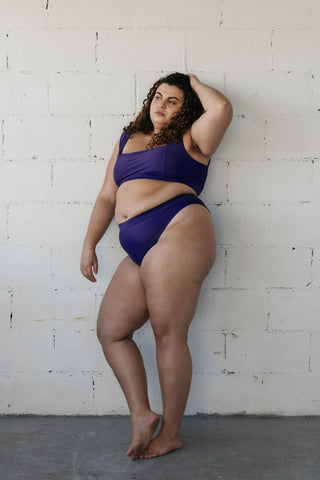 A woman leaning against a white brick wall with one hand in her hair wearing dark purple high waisted bikini bottoms and a matching dark purple supportive bikini top.