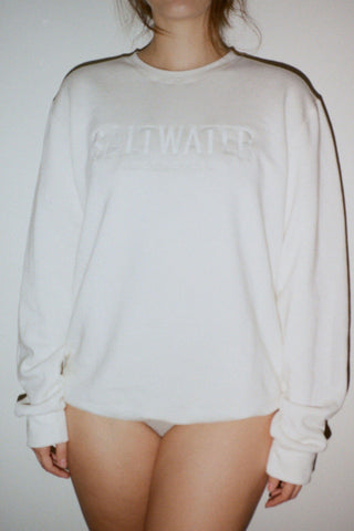 A woman wearing an oversized white long sleeve sweatshirt with a logo across the front. 