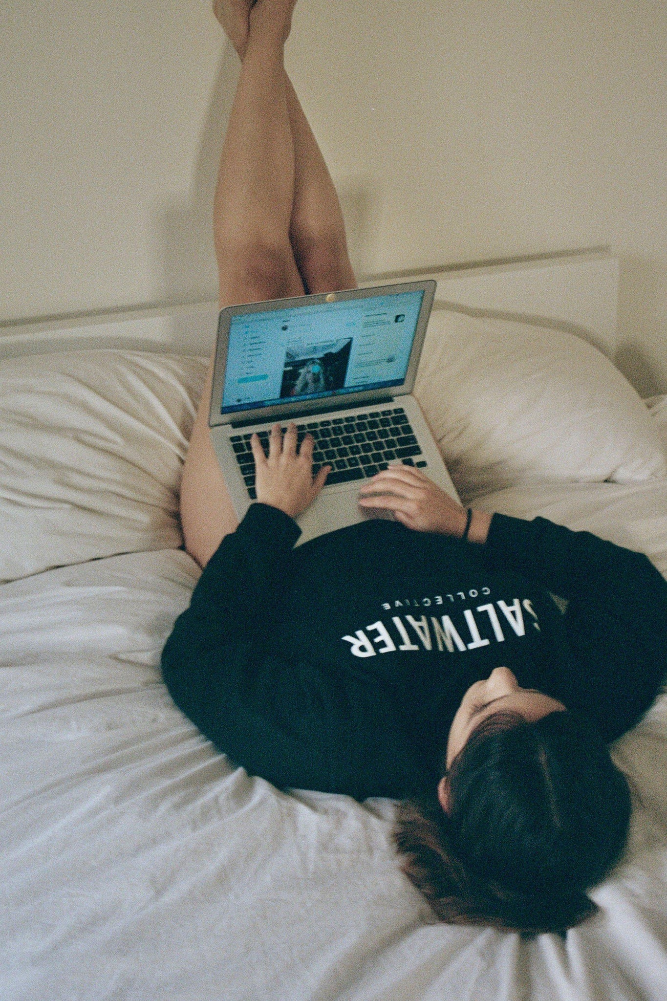 A woman laying on a bed with her legs leaned up against the wall wearing a black crewneck sweatshirt.