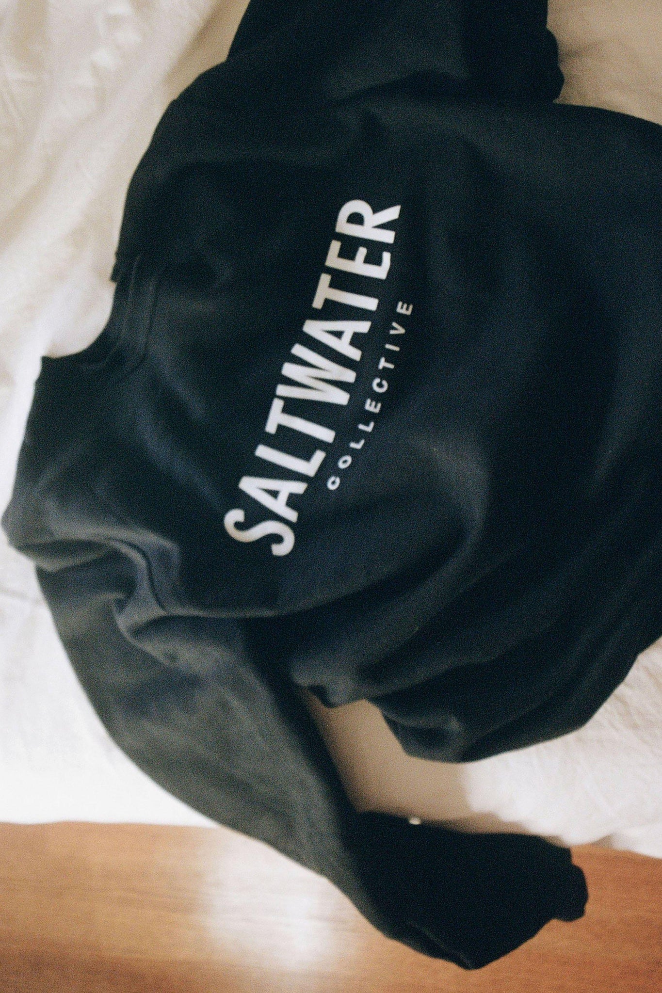 A black sweatshirt laying on a white bed with a white bold logo printed across the chest.