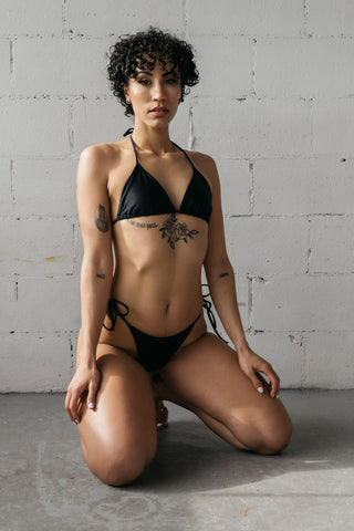 Woman kneeling on the ground with her hands on her thighs wearing black adjustable triangle bikini bottoms with a matching black string triangle bikini top.