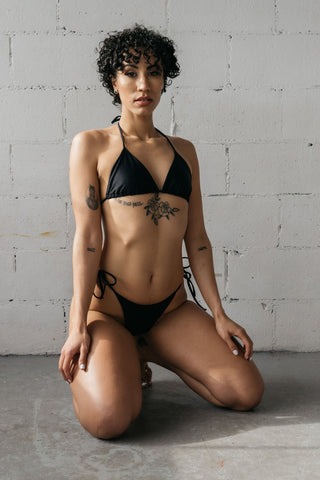 A woman kneeling on the ground wearing adjustable black triangle string bikini bottoms with a matching black triangle string bikini top.