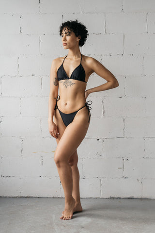 Woman standing with one hand on her hip wearing black adjustable triangle bikini bottoms with a matching black string triangle bikini top.