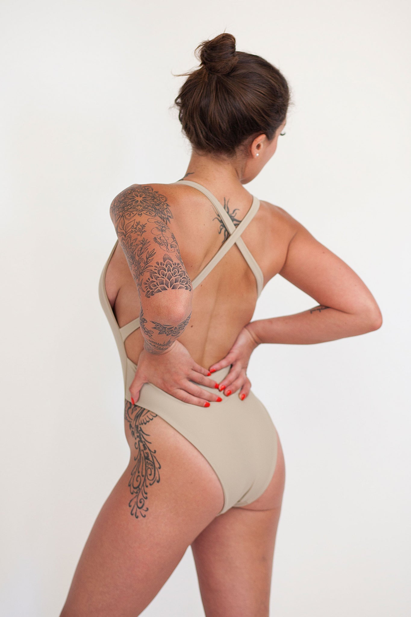 The back of a woman standing with her hands on her hips wearing a nude one piece swimsuit with criss-crossed straps.