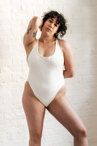 A woman standing in front of a white brick wall with one arm above her head and the other behind her back wearing a white high cut one-piece swimsuit.