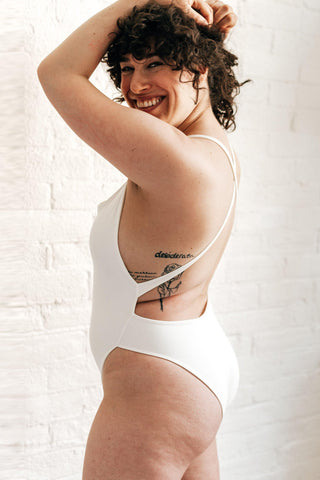 A woman standing to the side and smiling over her shoulder wearing a white one piece swimsuit with criss-crossed straps.