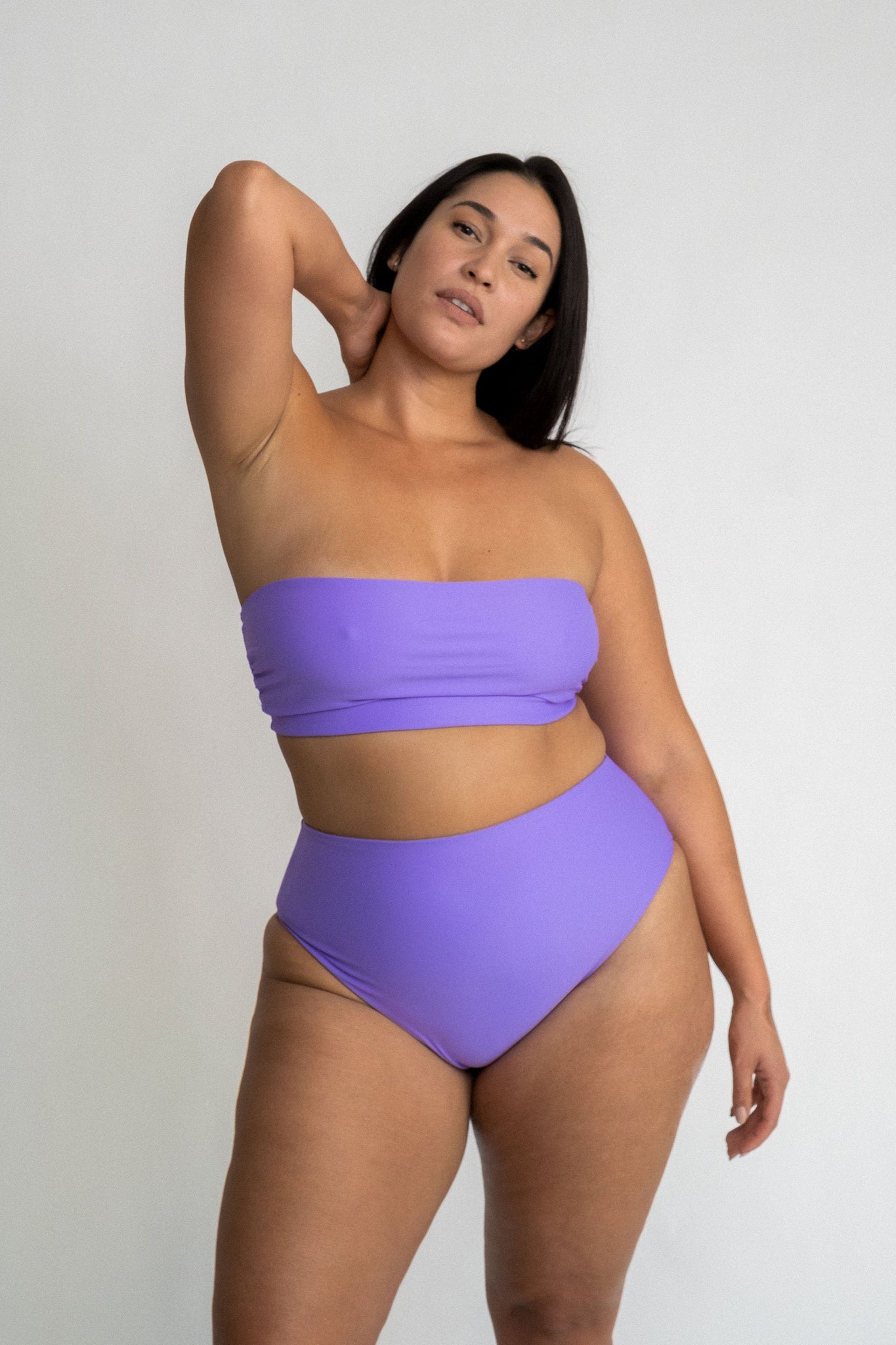 Woman standing with one arm above her head wearing high waisted bright purple bikini bottoms with a matching bright purple bandeau bikini top.