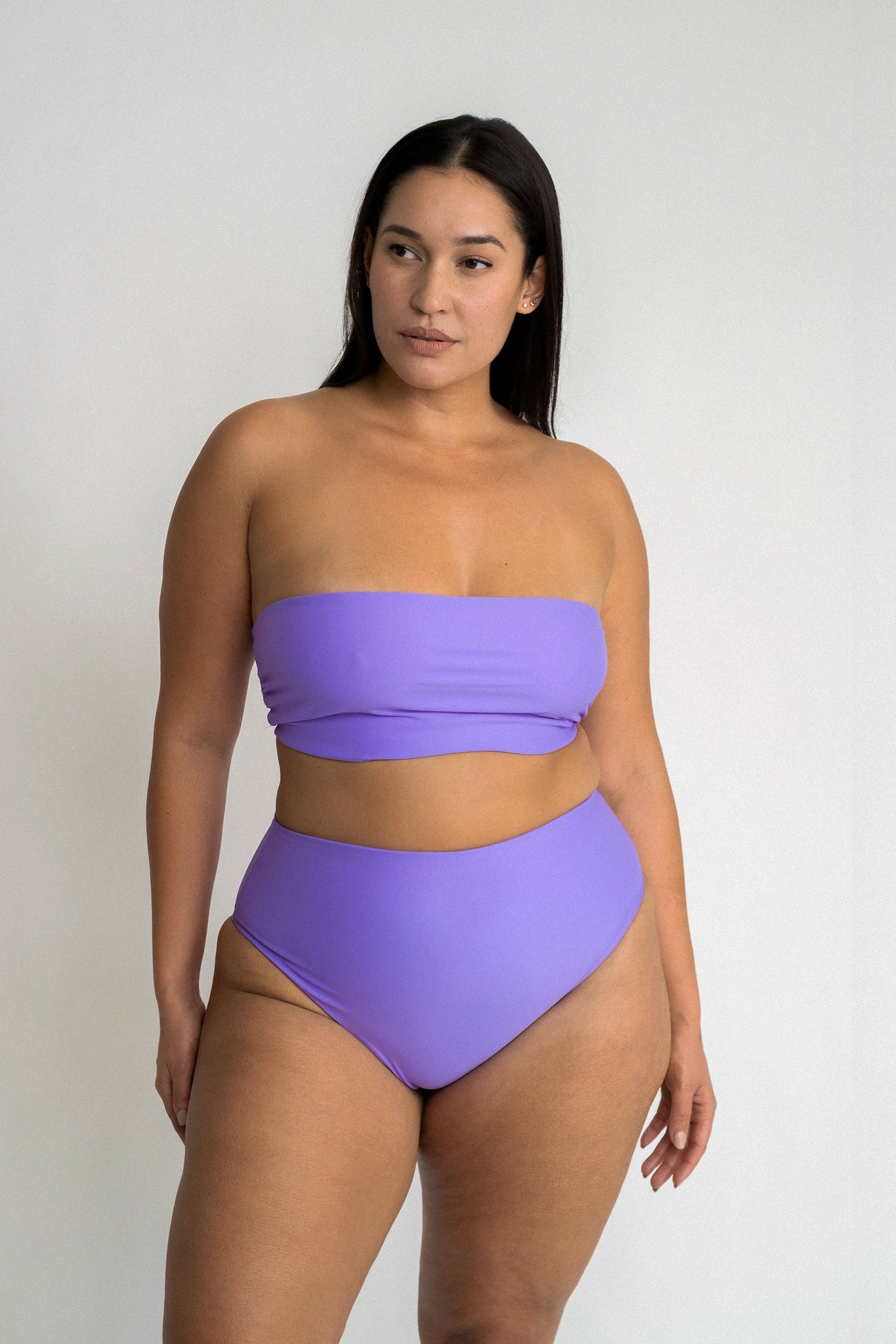 Woman standing in front of a white wall wearing bright purple high waisted bikini bottoms with a matching bright purple bandeau bikini top.