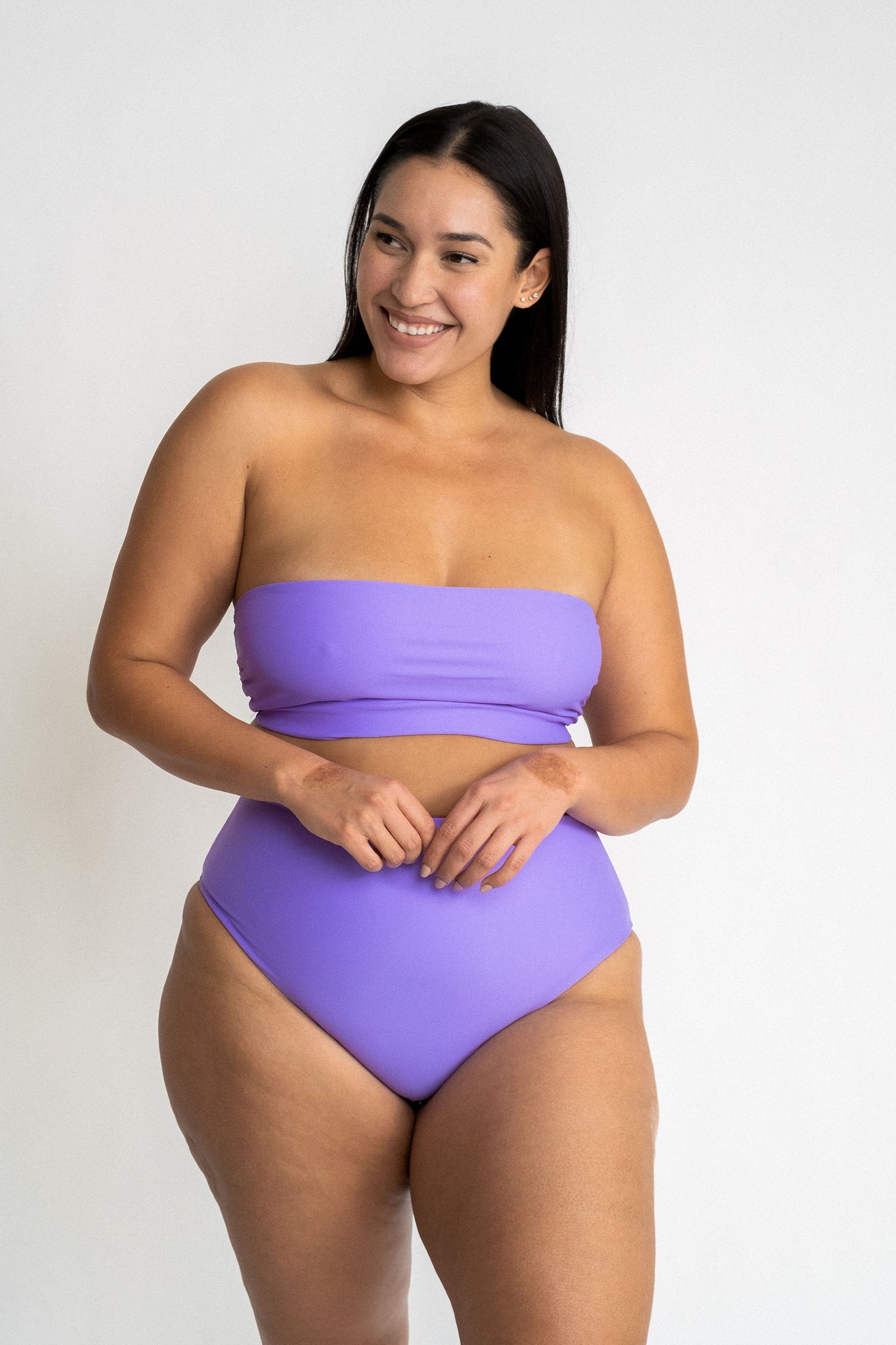 Woman standing with her hands crossed in front of her wearing bright purple high waisted bikini bottoms with a matching bright purple strapless bandeau bikini top.