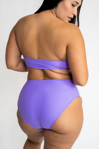 The back of a woman standing and looking over her shoulder wearing bright purple high waisted bikini bottoms with a matching bright purple strapless bandeau bikini top.