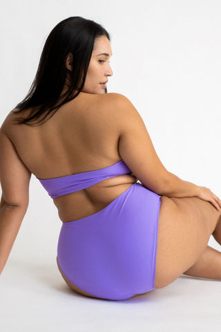 Woman sitting on the ground leaning against one arm wearing bright purple high waisted bikini bottoms with a matching bright purple strapless bandeau bikini top.