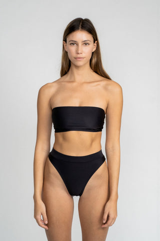 A woman standing with her arms by her side in front of a white wall wearing black high waisted bikini bottoms with a matching black strapless bandeau bikini top.