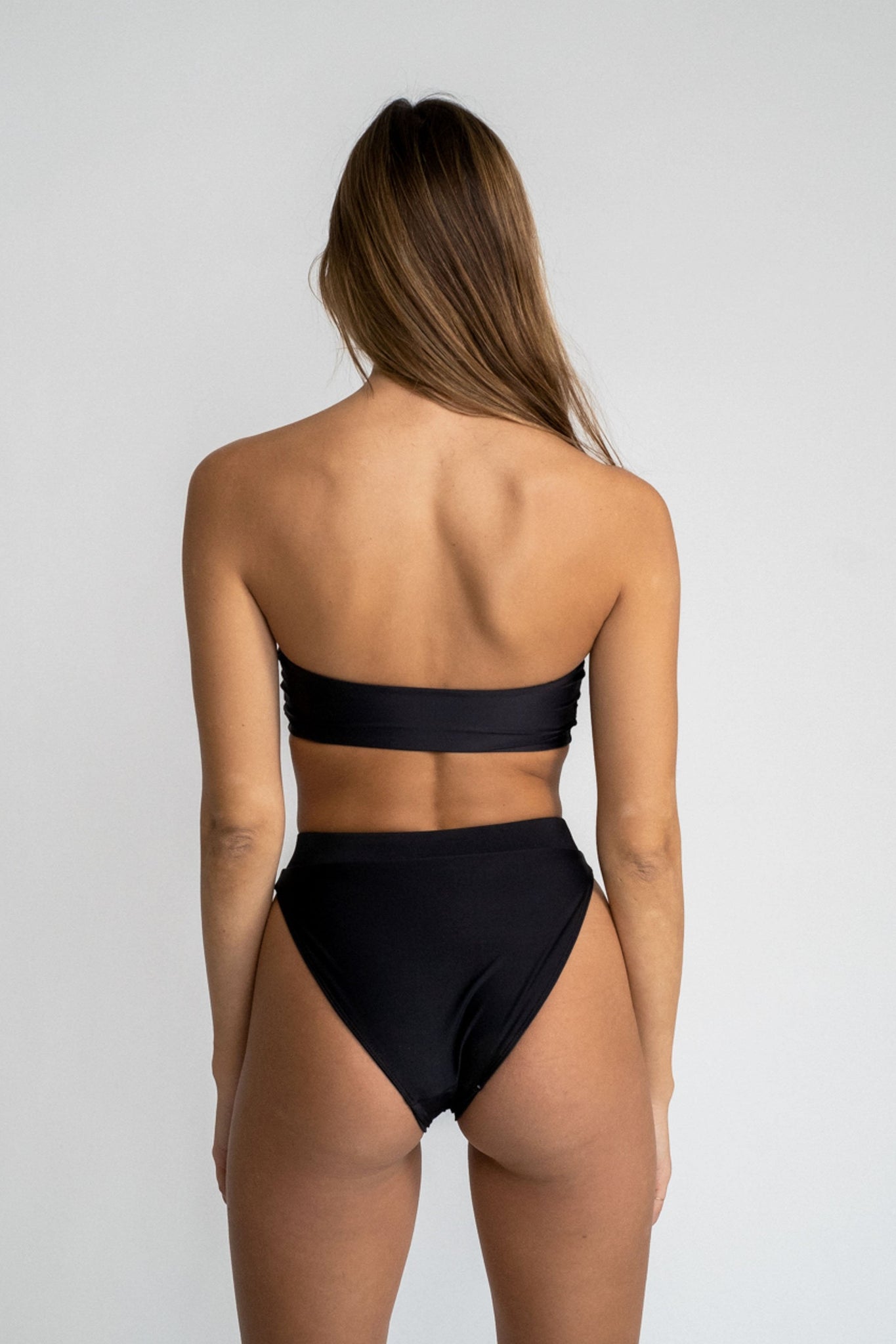 The back of a woman standing in front of a white wall wearing black high waisted bikini bottoms with a matching black strapless bandeau top.