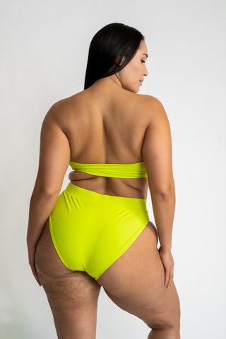 The back of a woman looking to the side standing in front of a white wall wearing bright neon green high waisted bikini bottoms with a matching bright green strapless bandeau bikini top.