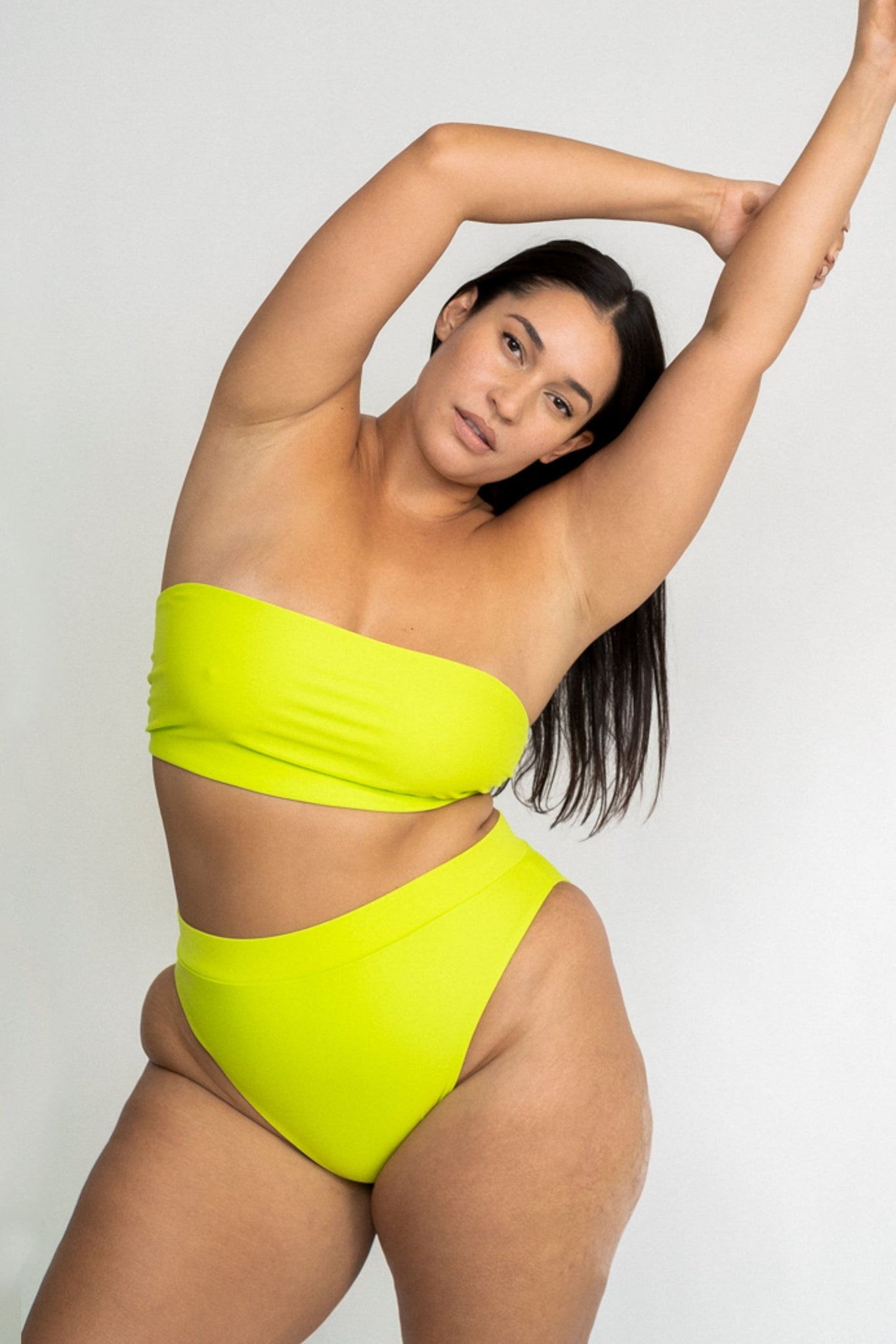 A woman leaning to the side with her arms above her head wearing bright neon green high waisted bikini bottoms with a matching bright neon green strapless bandeau bikini top.