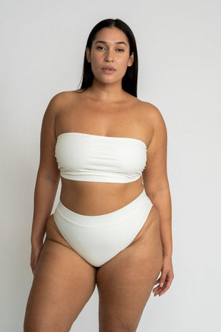 A woman standing in front of a white wall with her arms by her side wearing white high waisted bikini bottoms with a matching white strapless bandeau bikini top.