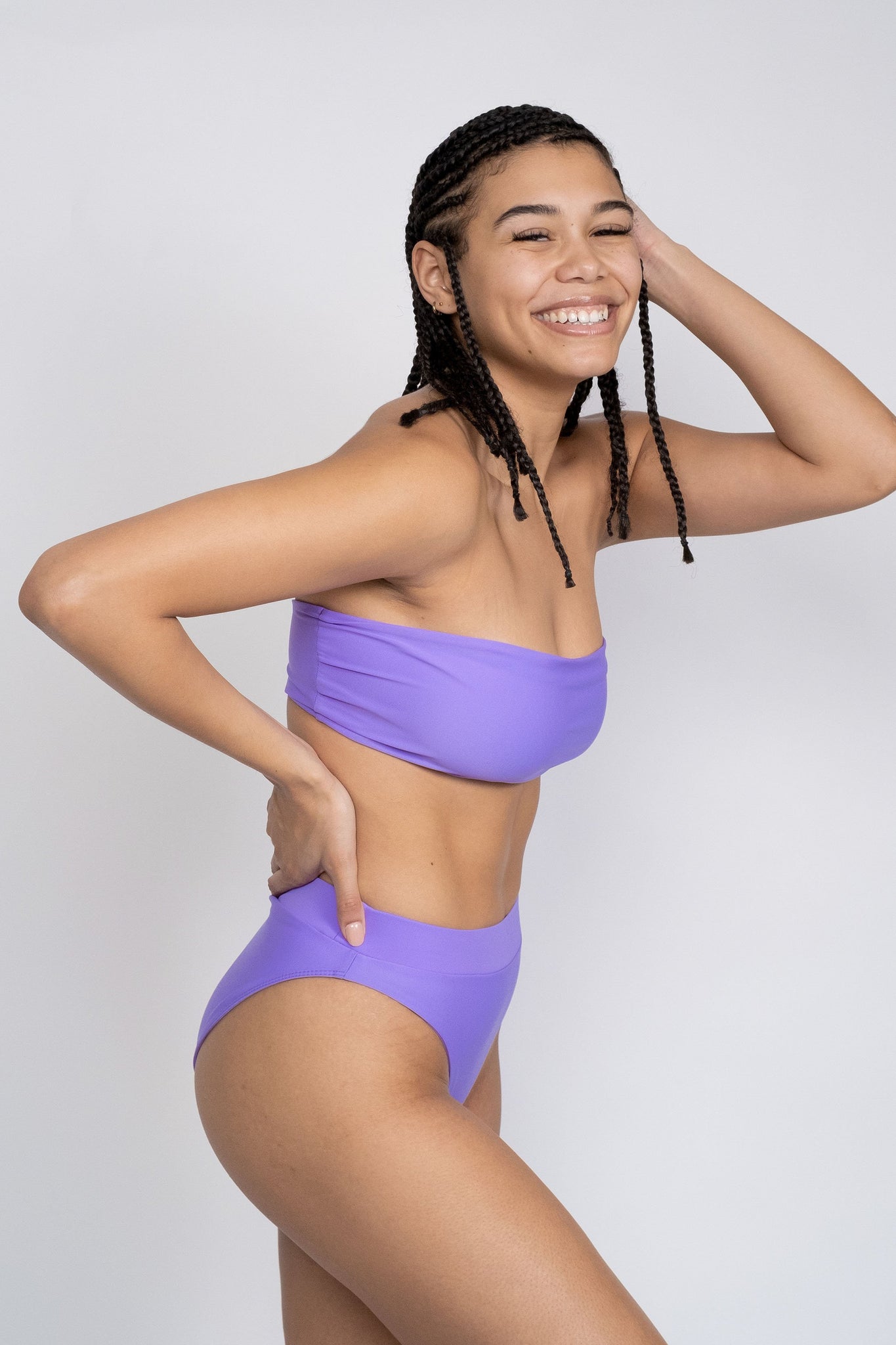 A woman laughing standing in front of a white wall with one arm in her hair and the other on her hip wearing bright purple high waisted bikini bottoms with a matching bright purple strapless bandeau top.