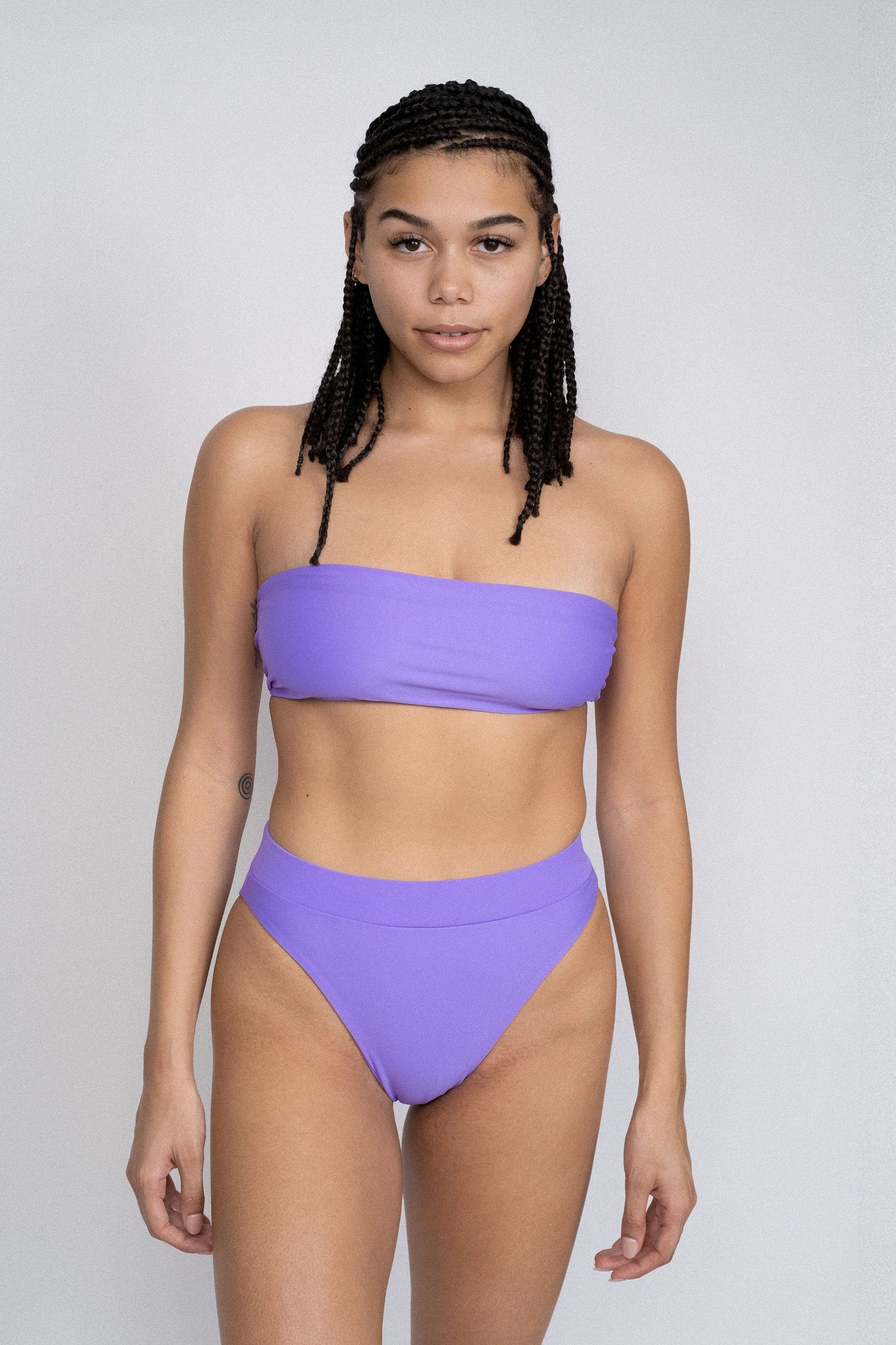 A woman standing with her arms by her side wearing bright purple high waisted bikini bottoms and a matching bright purple strapless bikini top.
