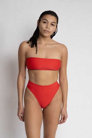 A woman standing in front of a white wall with her head leaned slightly to the side wearing bright red high waisted bikini bottoms with a matching bright red strapless bandeau bikini top.