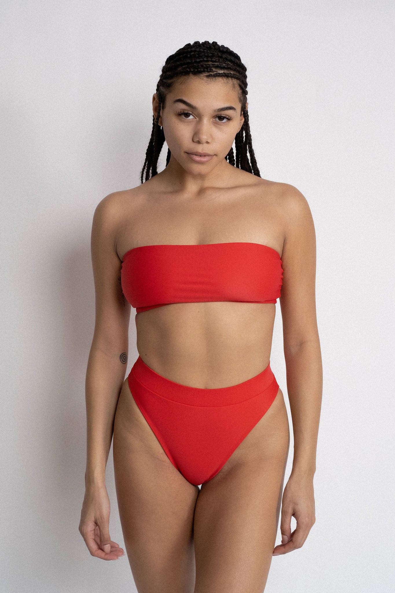 A woman standing in front of a white wall with her arms by her side wearing bright red high waisted bikini bottoms with a matching bright red strapless bandeau bikini top. 